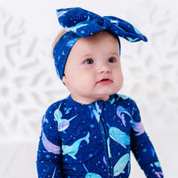 Close up portrait of a baby in a bow headband looking up, wearing a zip up pajama in a royal blue print of blue and lavender narwhals.