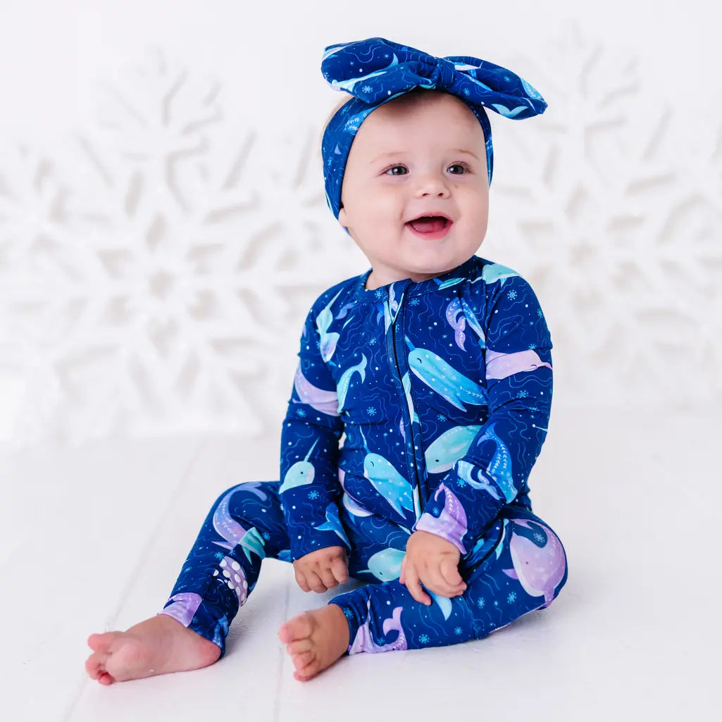 A smiling baby in a bow headband sitting barefoot on a white floor with hands resting in the lap, shown full length in a zip up pajama in a royal blue print of blue and lavender narwhals.