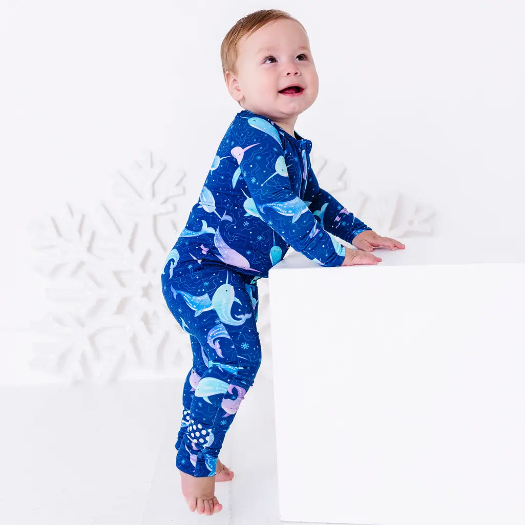 Side view of a smiling baby standing barefoot and leaning on a white block, in a zip up pajama with grip dots on the foot in a royal blue print of blue and lavender narwhals.