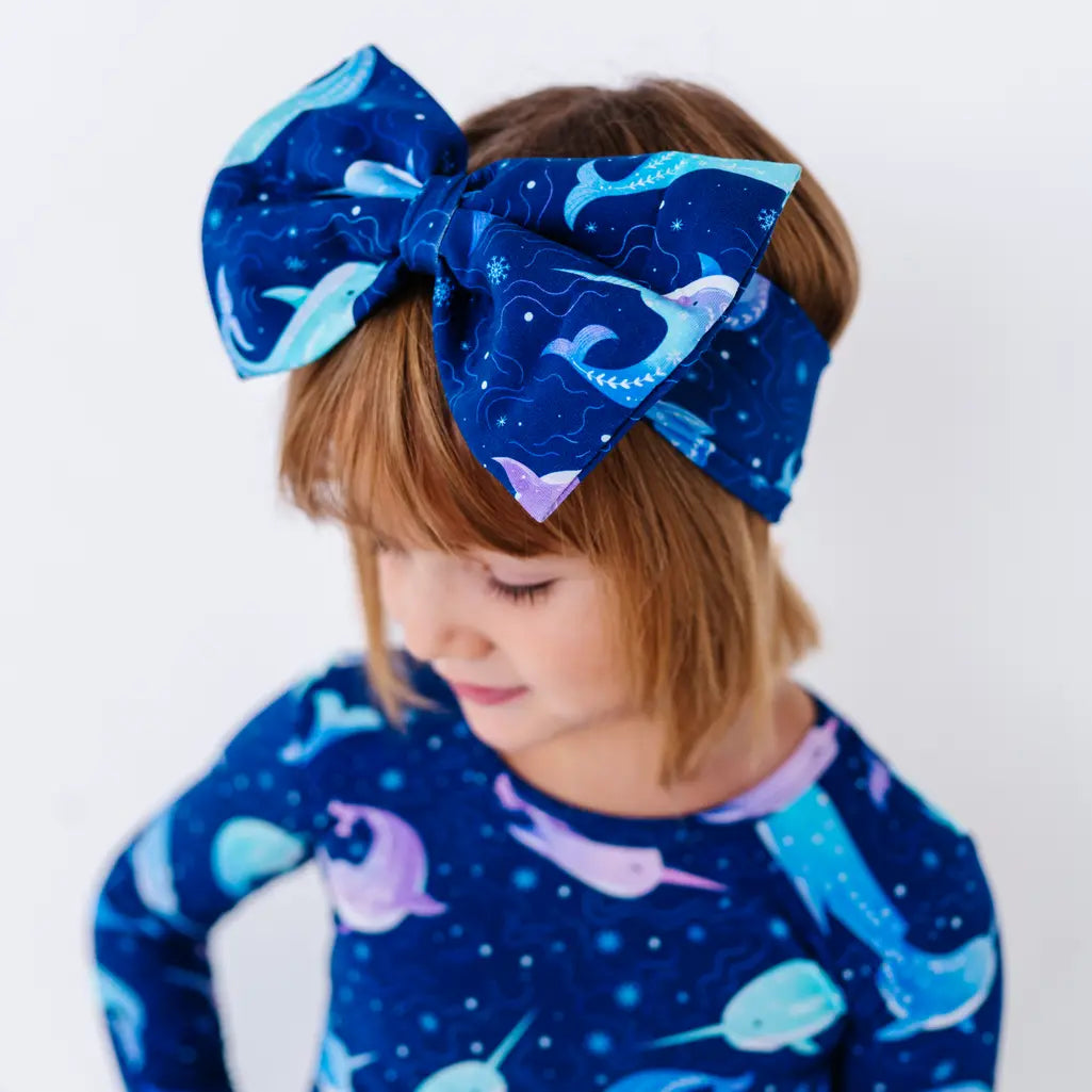 A child with short hair looking down to one side while wearing a large fabric bow headband in a royal blue print of blue and lavender narwhals, with a matching long sleeve top.
