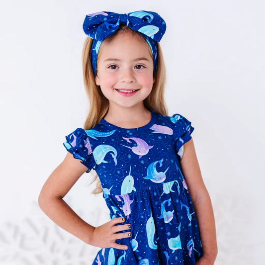 A smiling child with long blonde hair and hands on hips, wearing a large fabric bow headband in a royal blue print of blue and lavender narwhals, with a matching flutter sleeve top.