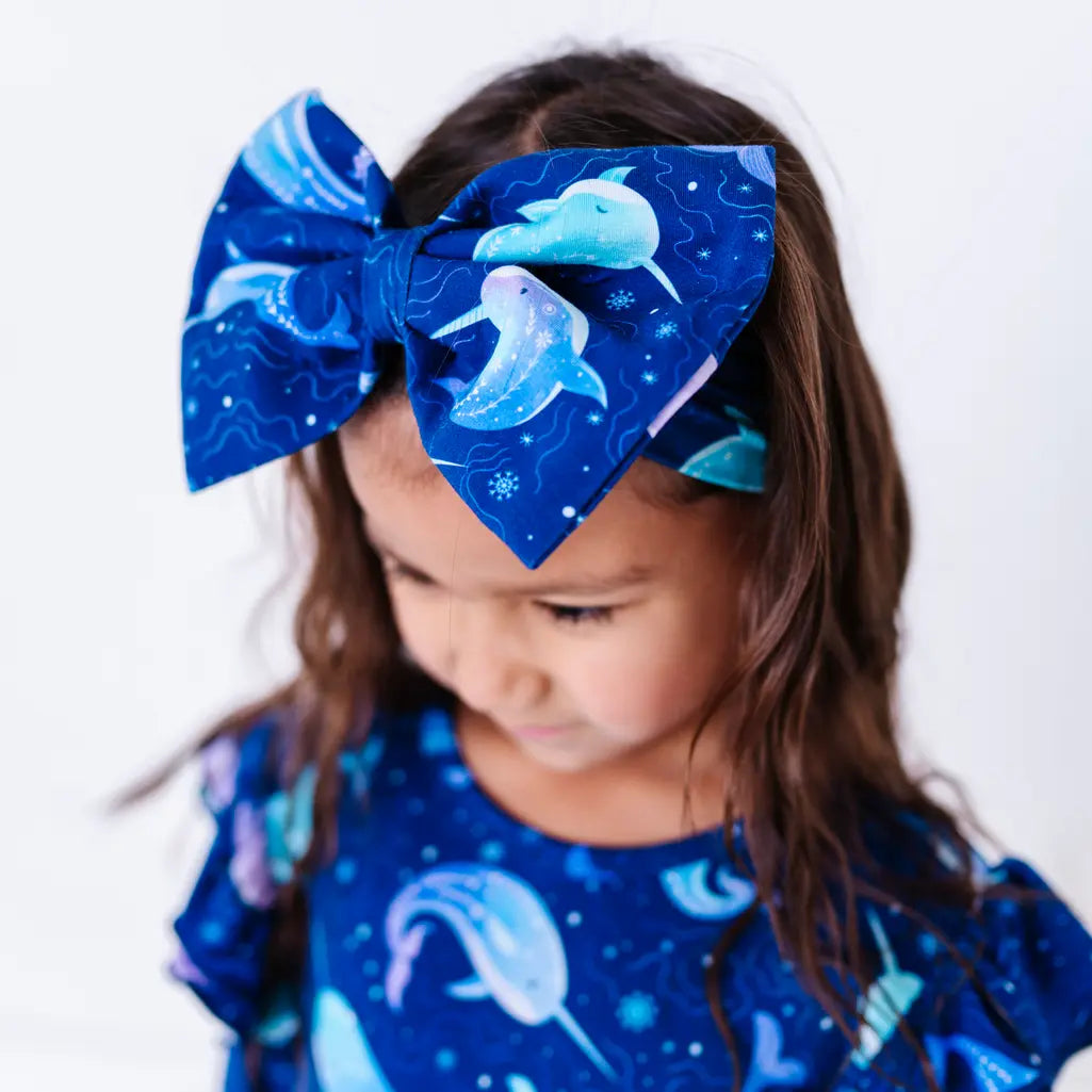 A child with long wavy hair looking down while wearing a large fabric bow headband in a royal blue print of blue and lavender narwhals, with a matching top.