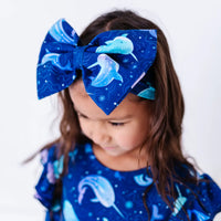 A child with long wavy hair looking down while wearing a large fabric bow headband in a royal blue print of blue and lavender narwhals, with a matching top.