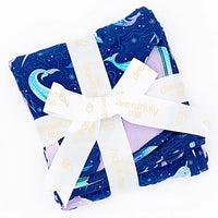 Flat lay of a folded royal blue blanket with a lavender reverse tied with a white ribbon in a print of blue and lavender narwhals on a white background.