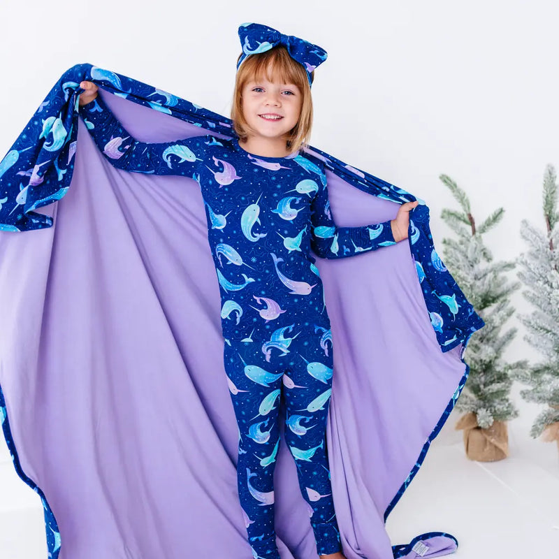 A smiling child in matching pajamas holding open a royal blue blanket with a lavender reverse in a print of blue and lavender narwhals like a cape.