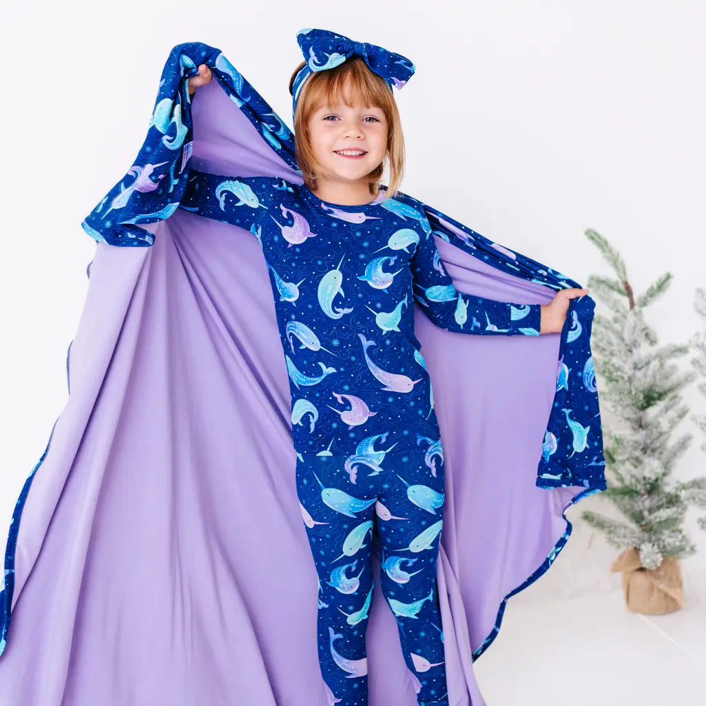 A smiling child with one arm raised, holding a wide royal blue blanket with a lavender reverse in a print of blue and lavender narwhals like a cape.