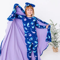 A smiling child with one arm raised, holding a wide royal blue blanket with a lavender reverse in a print of blue and lavender narwhals like a cape.