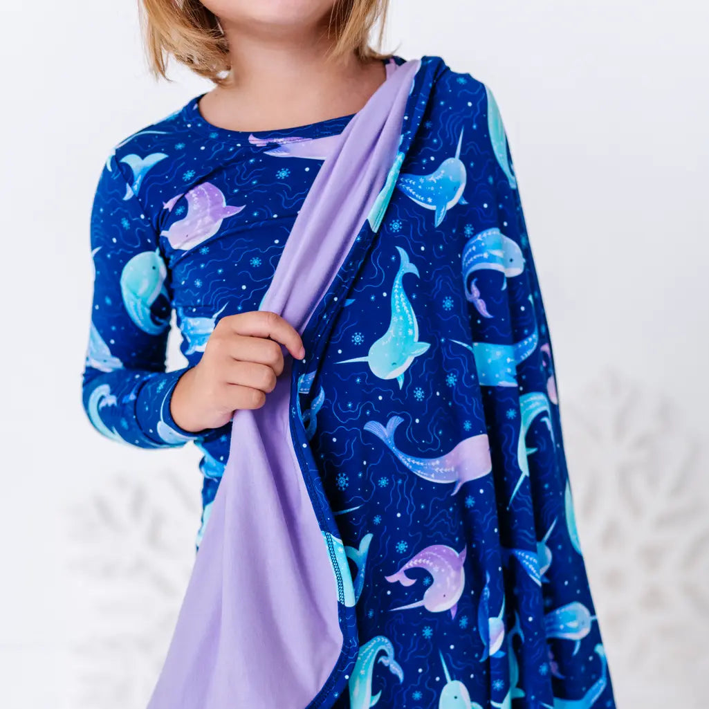Close up of a child holding the edge of a royal blue blanket with a lavender reverse in a print of blue and lavender narwhals draped over one shoulder.