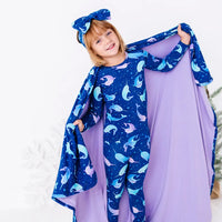 A child with the head tilted and a big smile, holding up the sides of a royal blue blanket with a lavender reverse in a print of blue and lavender narwhals like a cape.