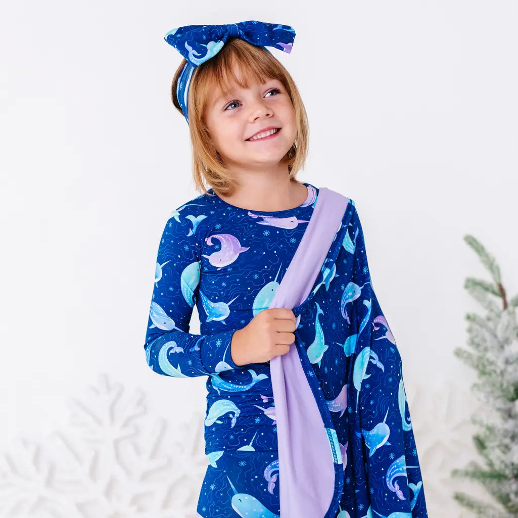 Portrait of a smiling child in a bow headband looking up, a royal blue blanket with a lavender reverse in a print of blue and lavender narwhals draped over one shoulder.