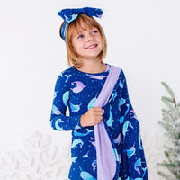 Portrait of a smiling child in a bow headband looking up, a royal blue blanket with a lavender reverse in a print of blue and lavender narwhals draped over one shoulder.