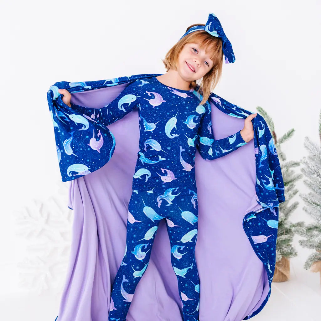 A child with the head tilted and arms stretched wide, holding out a royal blue blanket with a lavender reverse in a print of blue and lavender narwhals like wings.