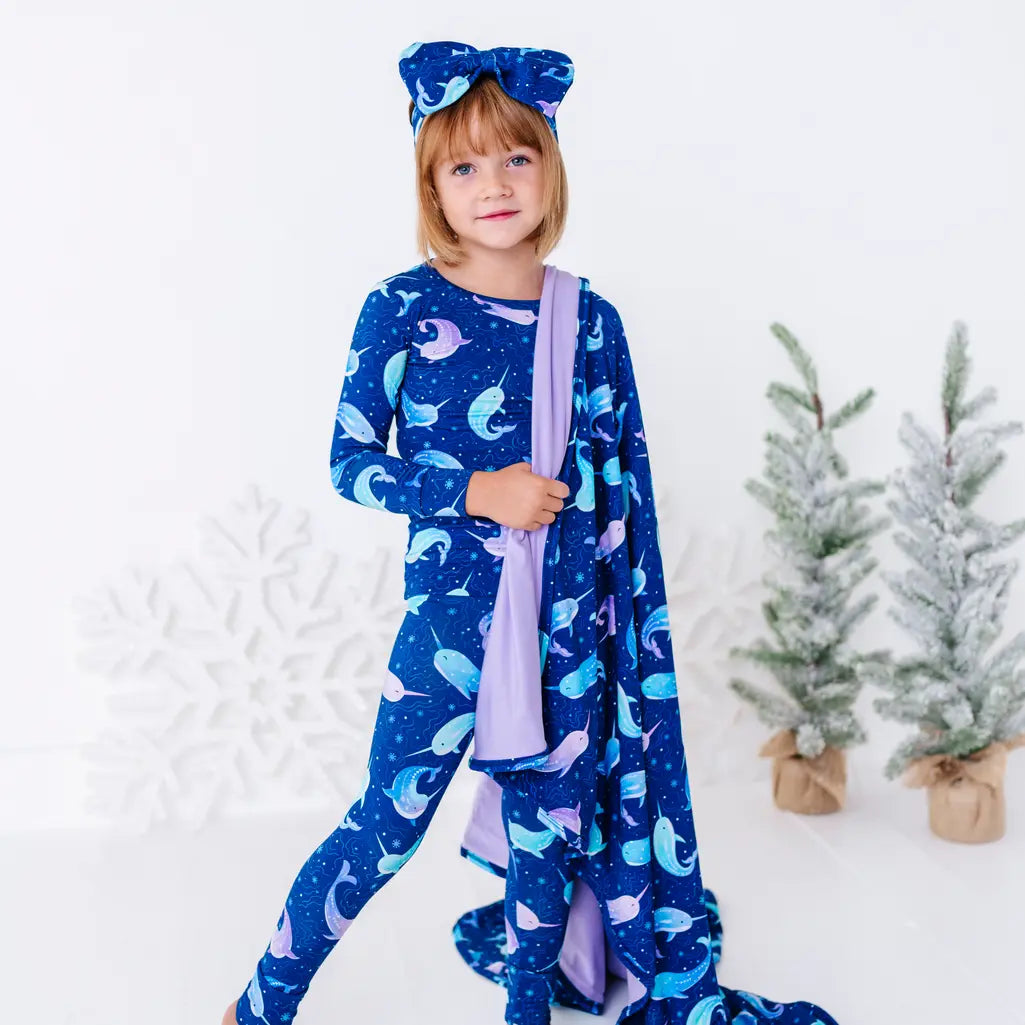 A child in matching pajamas standing mid step with a smile, a royal blue blanket with a lavender reverse in a print of blue and lavender narwhals draped over one arm and trailing to the floor.