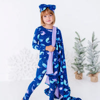 A child in matching pajamas standing mid step with a smile, a royal blue blanket with a lavender reverse in a print of blue and lavender narwhals draped over one arm and trailing to the floor.