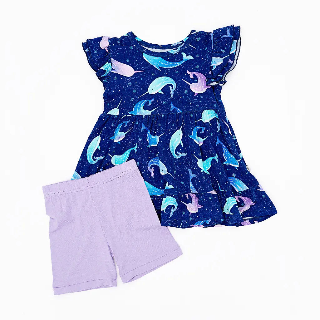 Flat lay of a flutter sleeve peplum top in a royal blue narwhal print with lavender bike shorts on a white background.