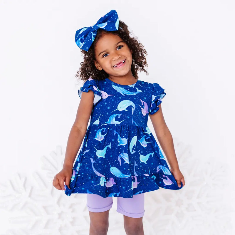 A child with a big smile holding out the hem of a flutter sleeve peplum top in a royal blue narwhal print, with lavender bike shorts.