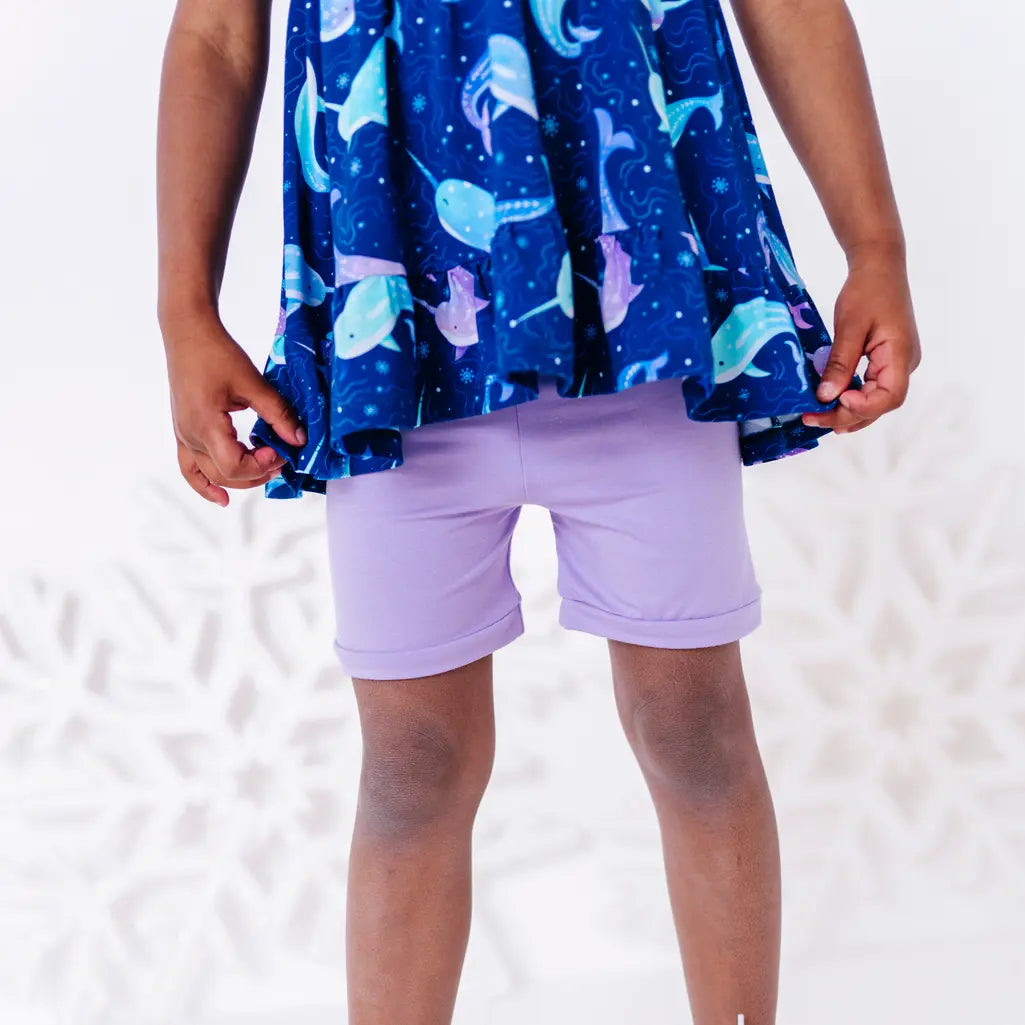 Close up of the ruffled hem of a flutter sleeve peplum top in a royal blue narwhal print above plain lavender bike shorts.