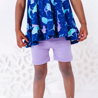 Close up of the ruffled hem of a flutter sleeve peplum top in a royal blue narwhal print above plain lavender bike shorts.