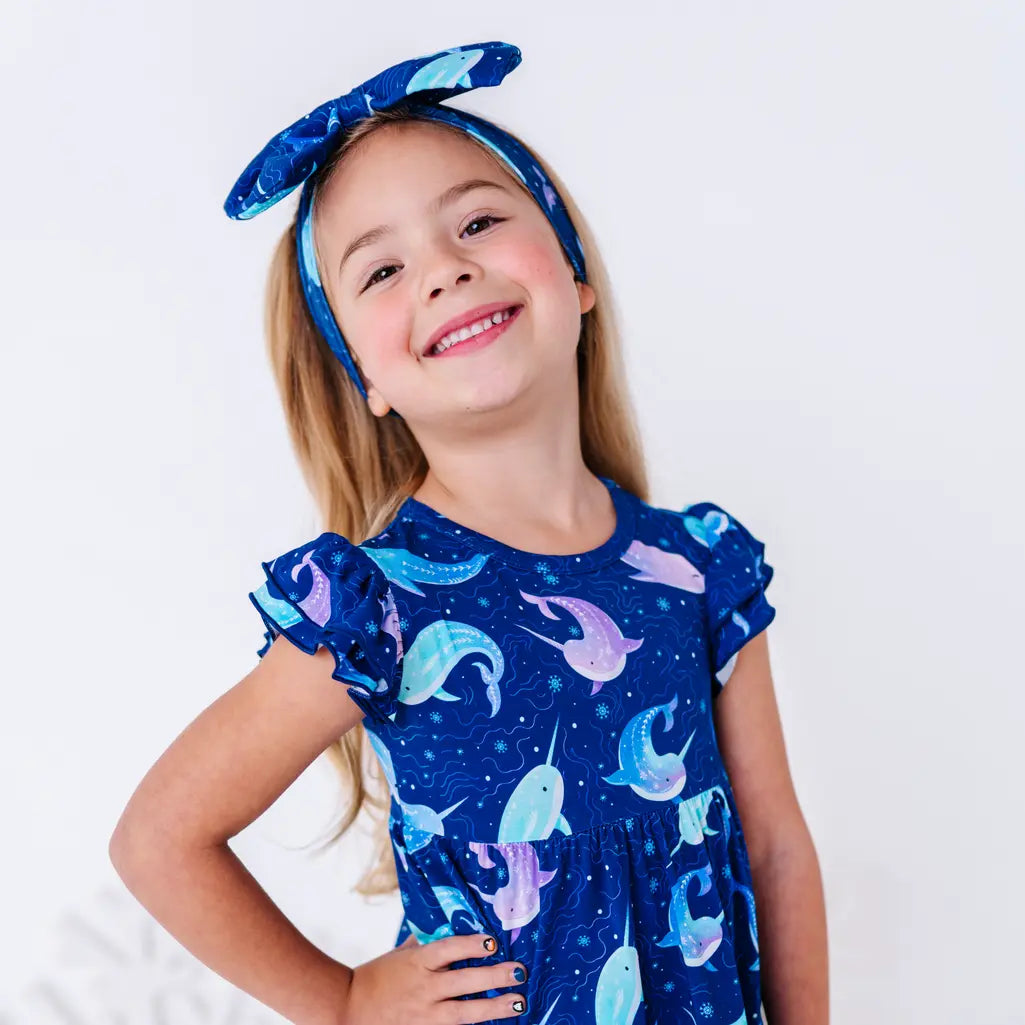 Close up portrait of a child with head tilted and a big smile, in a flutter sleeve peplum top in a royal blue narwhal print.