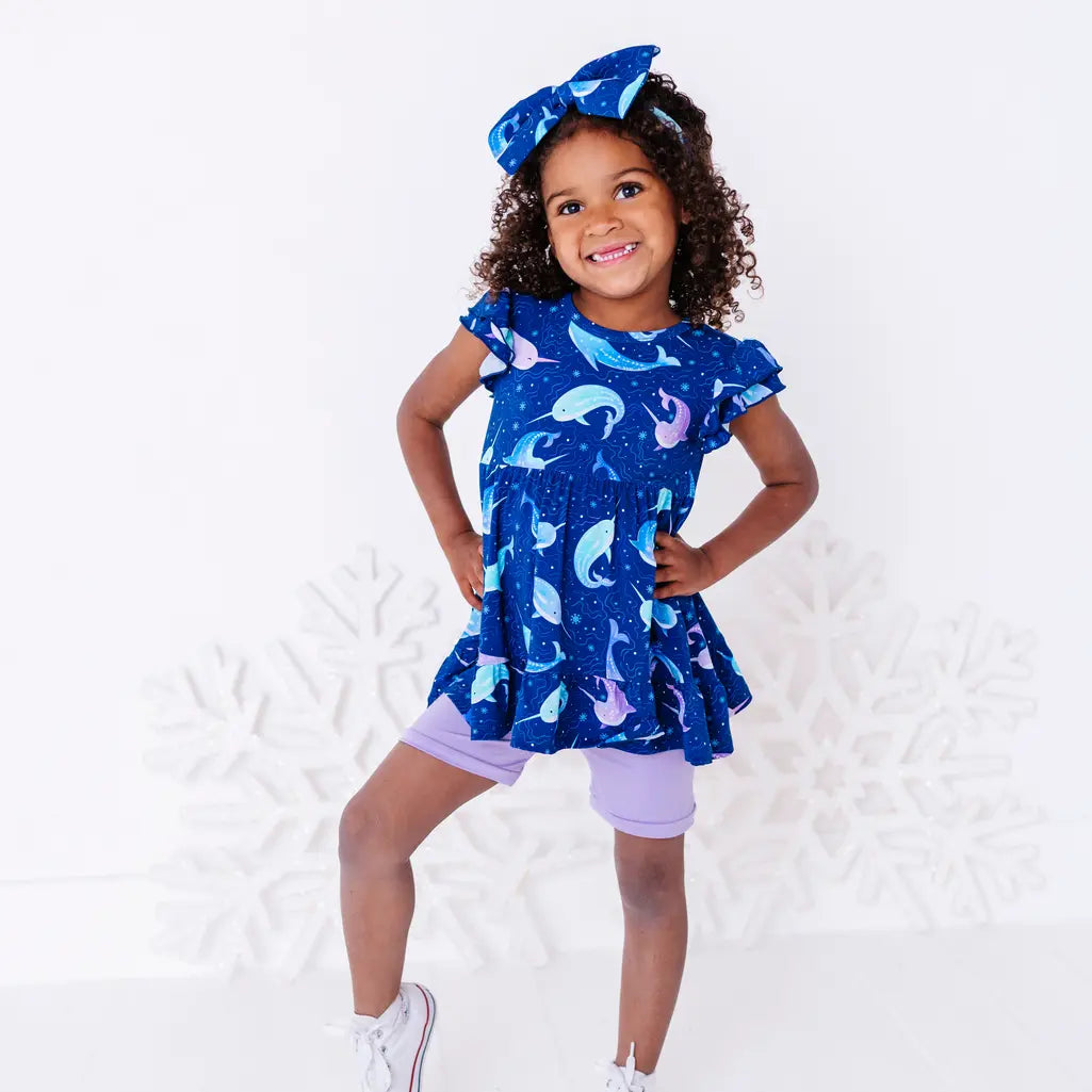 A child standing with hands on hips and one foot lifted, smiling, in a flutter sleeve peplum top in a royal blue narwhal print and lavender bike shorts.