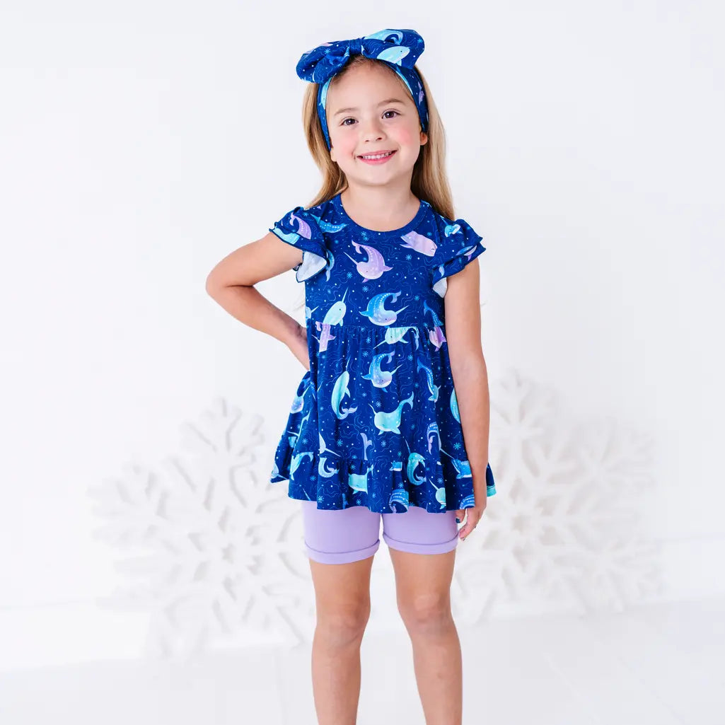 A child smiling with one hand on the hip, standing in a flutter sleeve peplum top in a royal blue narwhal print and lavender bike shorts.