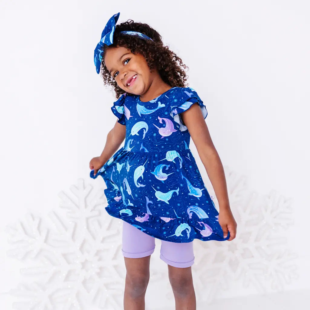 A child swaying with the hem of a flutter sleeve peplum top held out and the head tilted, in a royal blue narwhal print with lavender bike shorts.