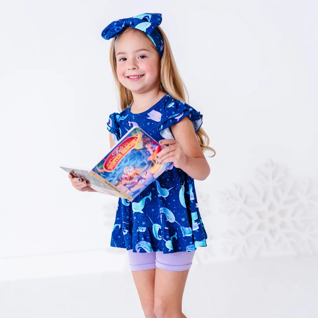 A child smiling at the camera while holding an open storybook, in a flutter sleeve peplum top in a royal blue narwhal print and lavender bike shorts.