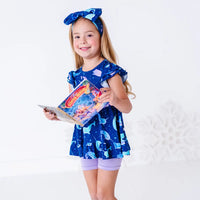 A child smiling at the camera while holding an open storybook, in a flutter sleeve peplum top in a royal blue narwhal print and lavender bike shorts.
