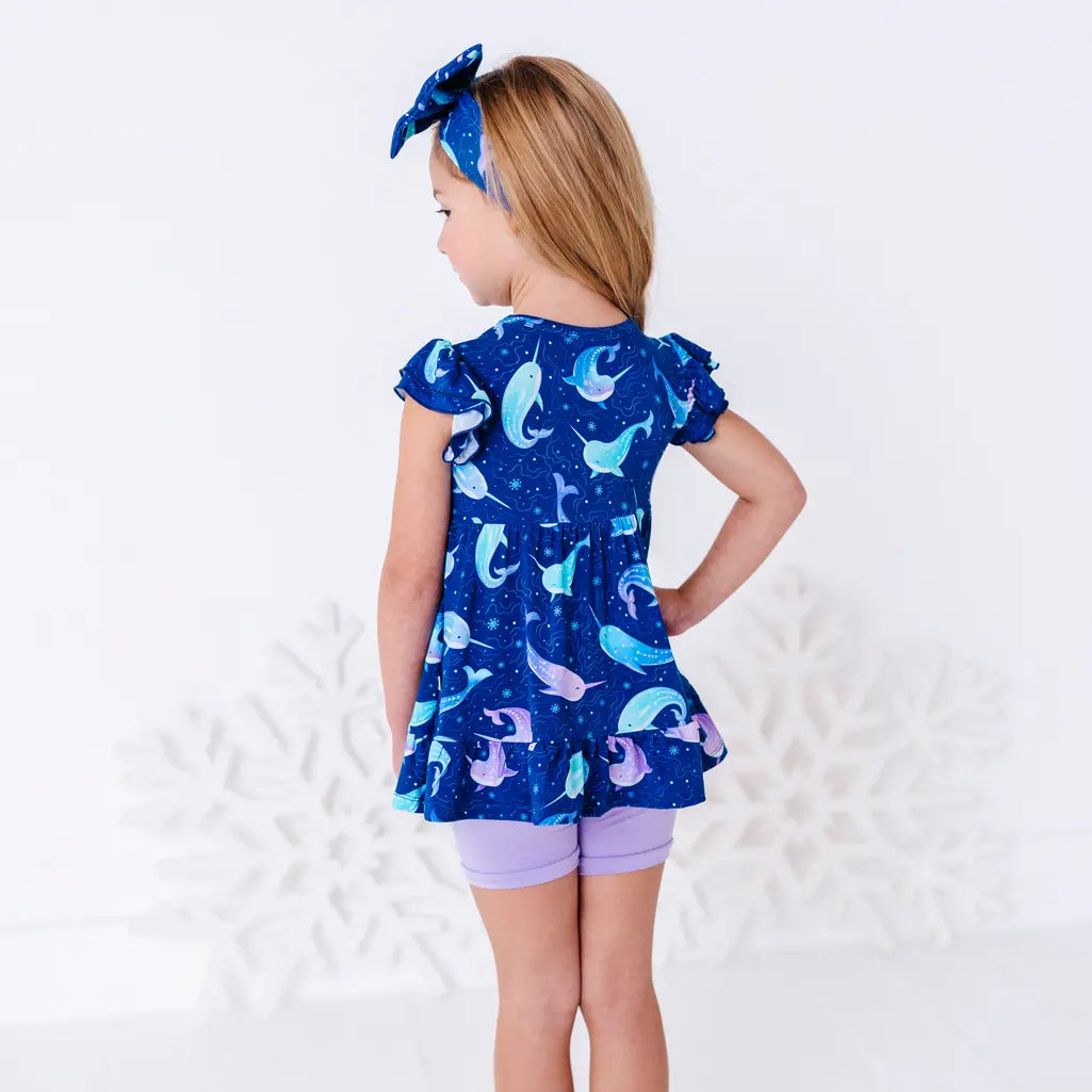 Back view of a child with the head turned to the side and a hand on the hip, in a flutter sleeve peplum top in a royal blue narwhal print and lavender bike shorts.