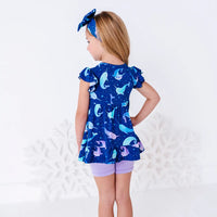Back view of a child with the head turned to the side and a hand on the hip, in a flutter sleeve peplum top in a royal blue narwhal print and lavender bike shorts.