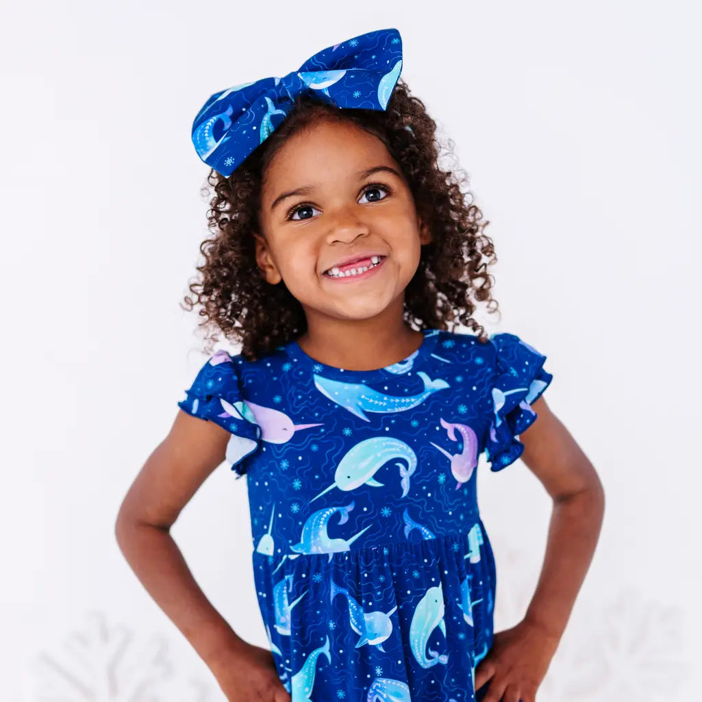 Close up portrait of a child with hands on hips and a smile, in a flutter sleeve peplum top in a royal blue narwhal print.