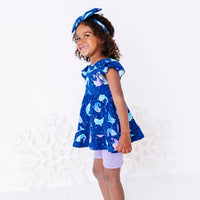 Side view of a child looking back at the camera with a smile, in a flutter sleeve peplum top in a royal blue narwhal print and lavender bike shorts.