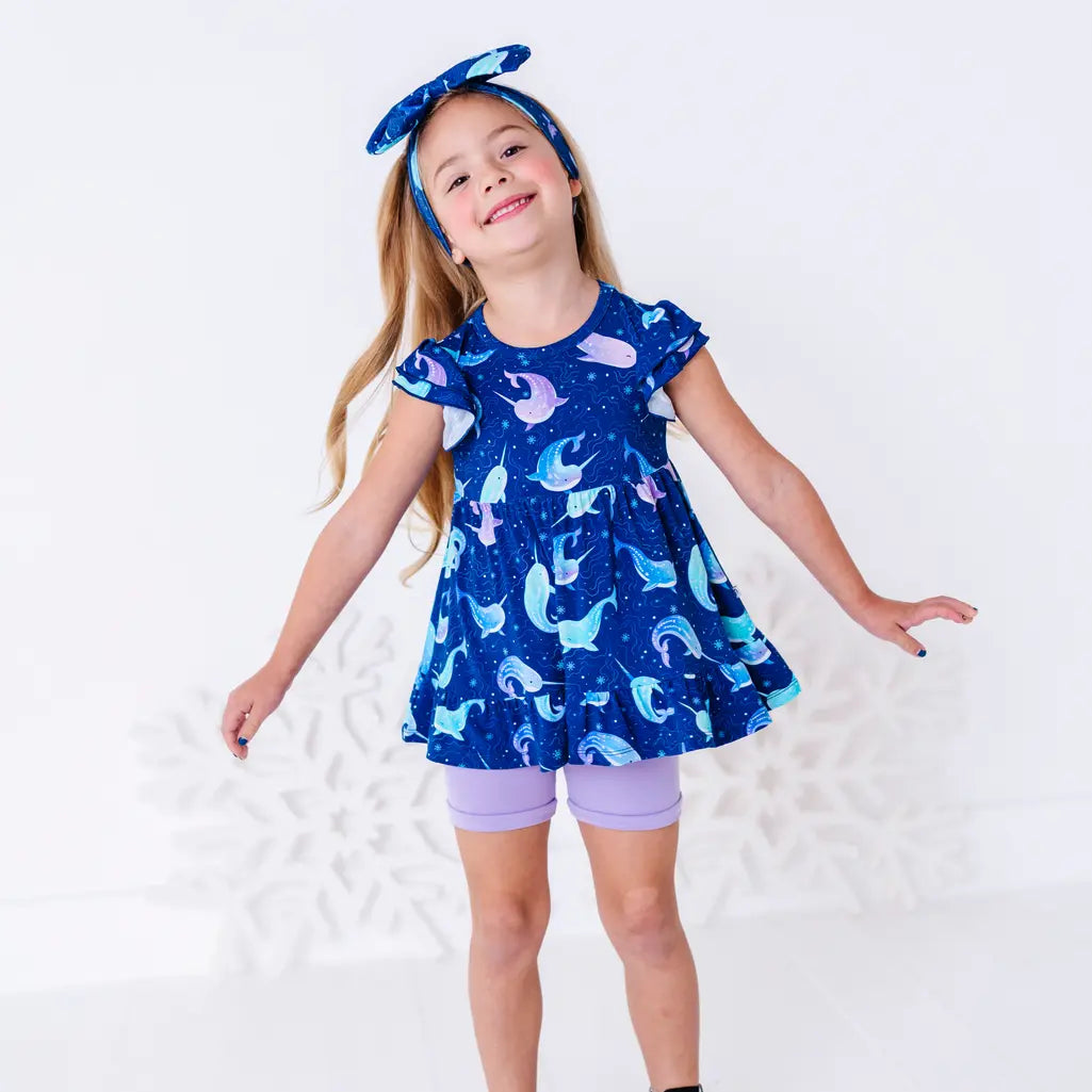 A child with arms out and a big smile, in a flutter sleeve peplum top in a royal blue narwhal print and lavender bike shorts.