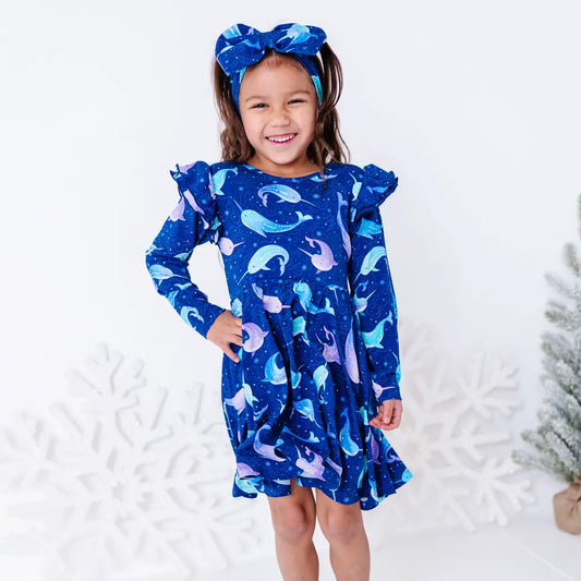 A child with a big smile and a hand on the hip, standing in a long sleeve twirl dress with flutter ruffles at the shoulders in a royal blue print of blue and lavender narwhals.