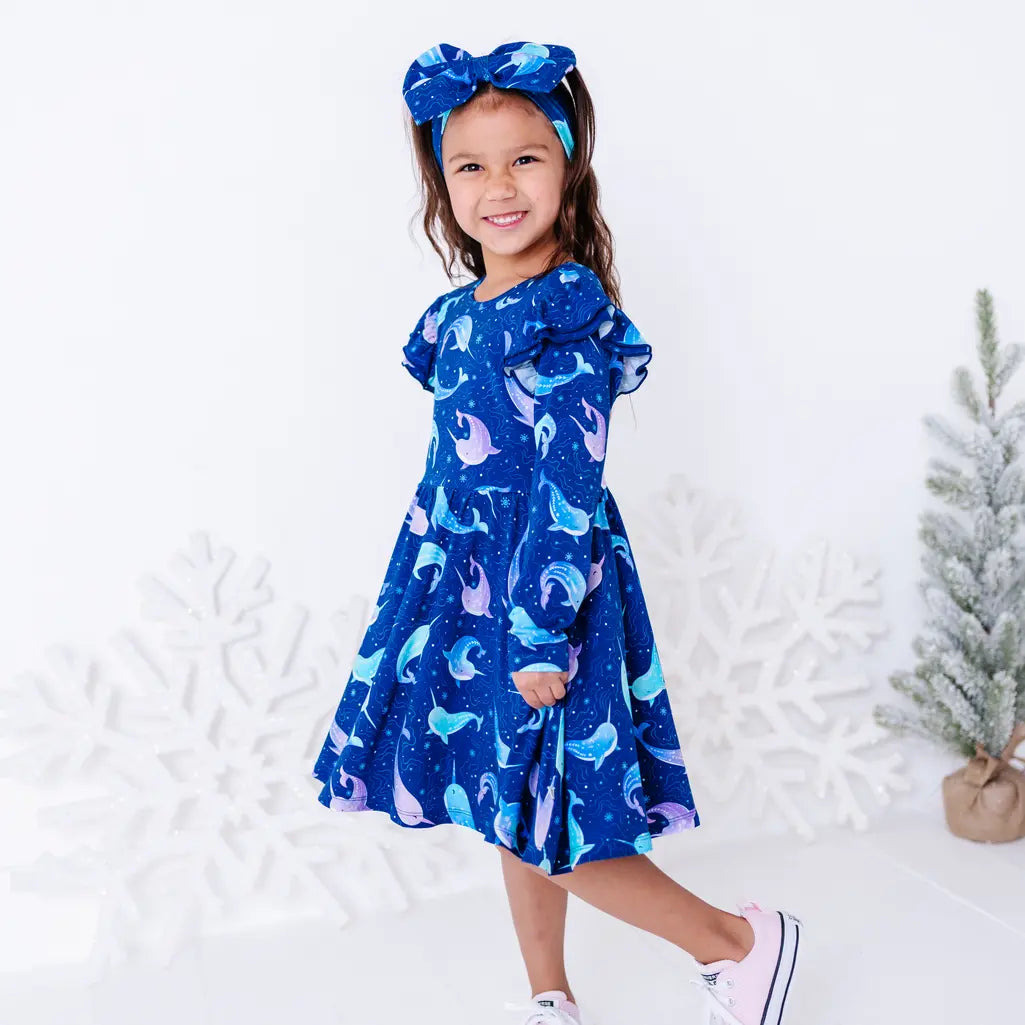 A child walking with a smile and the skirt of a long sleeve twirl dress in motion, with flutter ruffles at the shoulders in a royal blue print of blue and lavender narwhals.