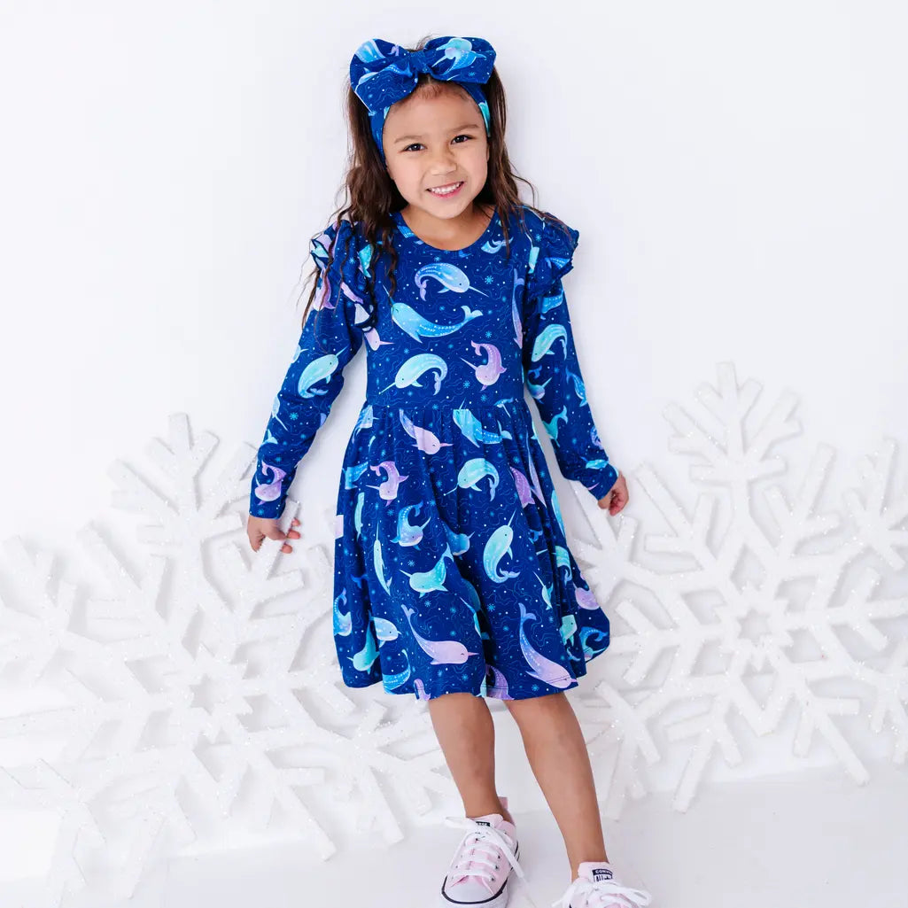A child smiling at the camera while standing full length with arms down, in a long sleeve twirl dress with flutter ruffles at the shoulders in a royal blue print of blue and lavender narwhals.