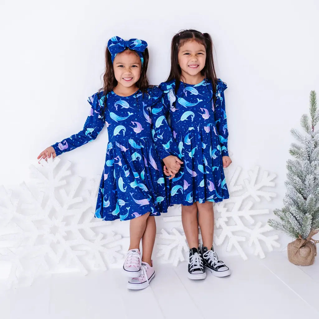 Two kids holding hands and smiling at the camera in matching long sleeve twirl dresses in a royal blue print of blue and lavender narwhals.