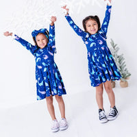 Two kids jumping with arms raised in matching long sleeve twirl dresses in a royal blue print of blue and lavender narwhals.