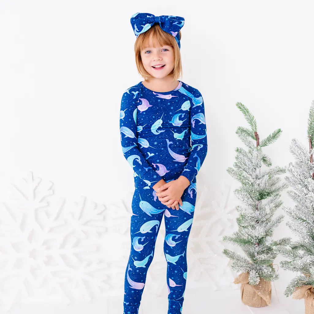 A child with a big smile and hands clasped, standing in a long sleeve pajama top and matching pants in a royal blue print of blue and lavender narwhals.