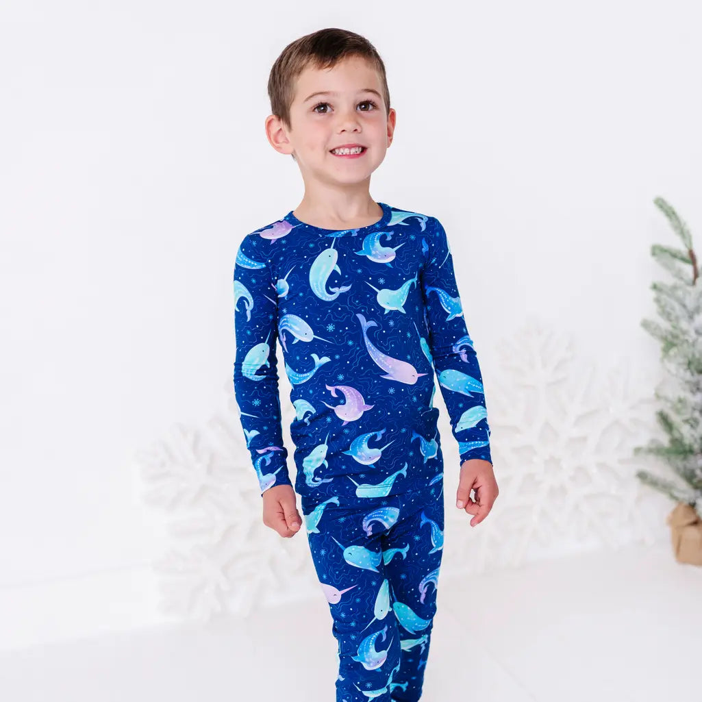 A child smiling at the camera and standing with arms relaxed, in a long sleeve pajama top and matching pants in a royal blue print of blue and lavender narwhals.