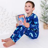 A child sitting barefoot on a white floor with a storybook and a big smile, in a long sleeve pajama top and matching pants in a royal blue print of blue and lavender narwhals.