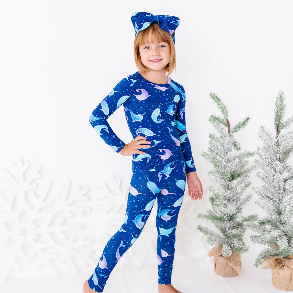 A child standing with one hand on the hip and smiling, in a long sleeve pajama top and matching pants in a royal blue print of blue and lavender narwhals.