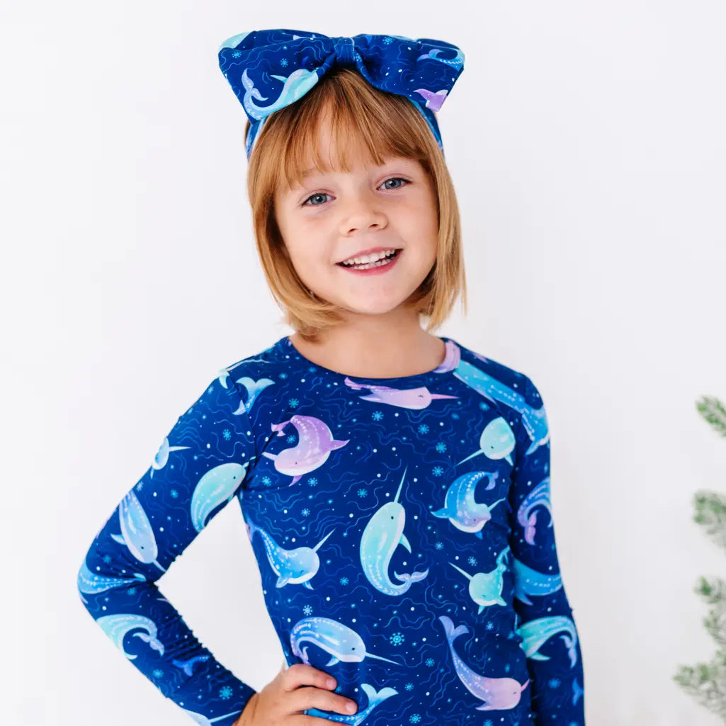 Close up portrait of a child with the head tilted and a hand on the hip, smiling in a long sleeve pajama top in a royal blue print of blue and lavender narwhals.