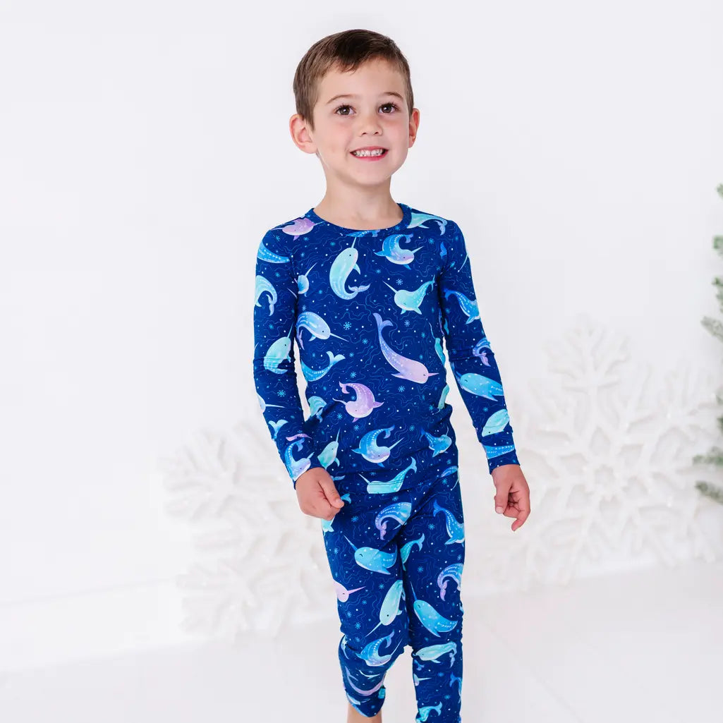 A child stepping forward with a smile, in a long sleeve pajama top and matching pants in a royal blue print of blue and lavender narwhals.