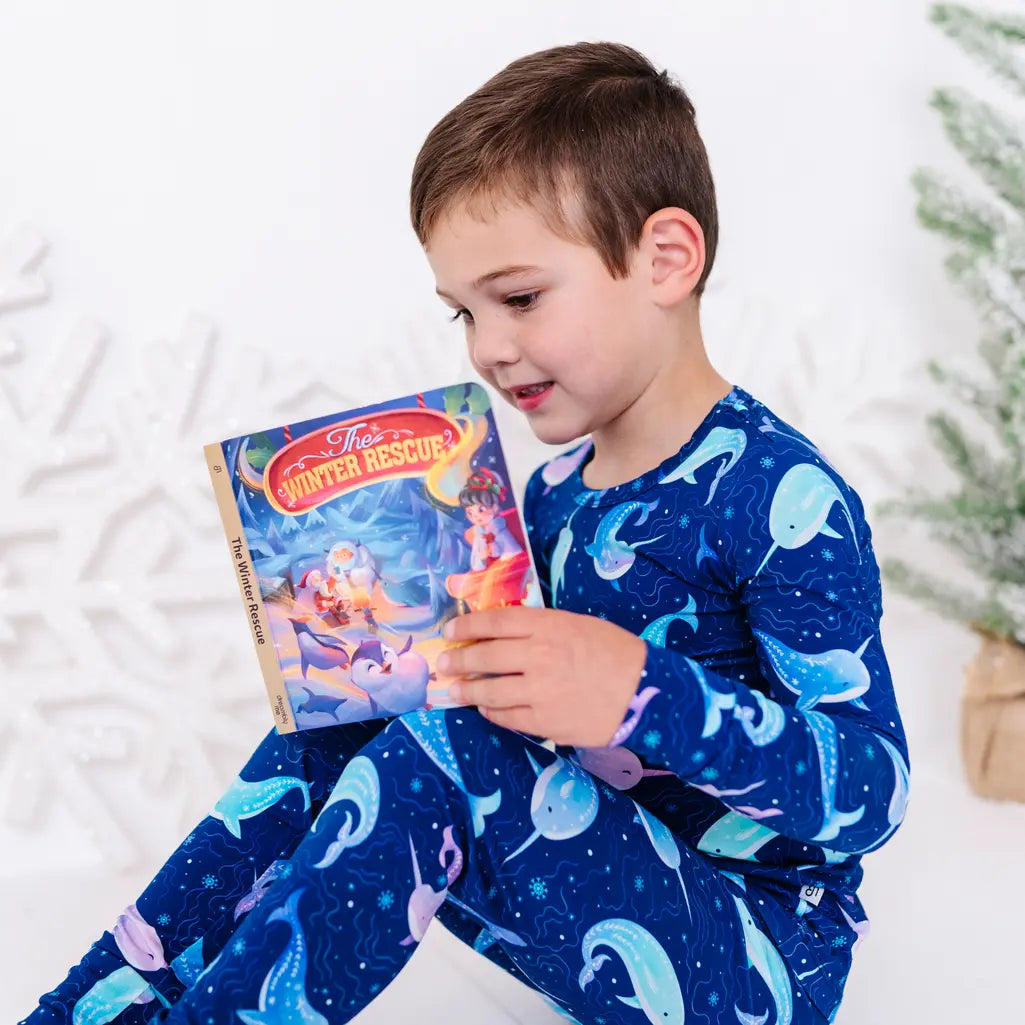 Side view of a child sitting on a white floor and reading a storybook, in a long sleeve pajama top and matching pants in a royal blue print of blue and lavender narwhals.