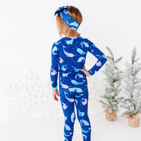 Back view of a child with the head turned to the side and a hand on the hip, in a long sleeve pajama top and matching pants in a royal blue print of blue and lavender narwhals.