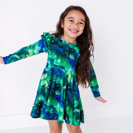 A child smiling at the camera with arms out and the skirt swinging, in a long sleeve twirl dress with flutter ruffles at the shoulders in a green and blue northern lights print with tiny stars.