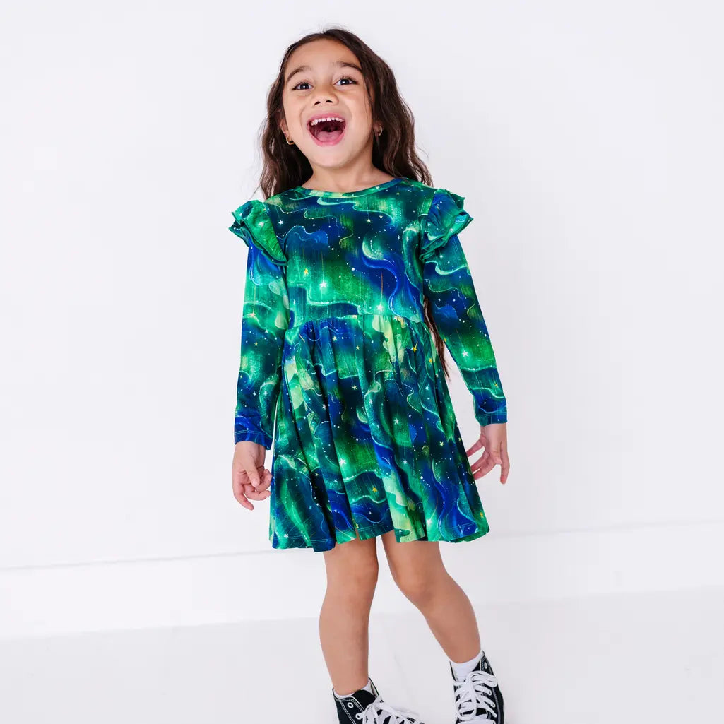 A child laughing with an open mouth and arms relaxed, standing in a long sleeve twirl dress with flutter ruffles at the shoulders in a green and blue northern lights print with tiny stars.