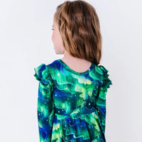 Close up of the back of a child with the head turned, showing the flutter ruffles on both shoulders of a long sleeve twirl dress in a green and blue northern lights print with tiny stars.