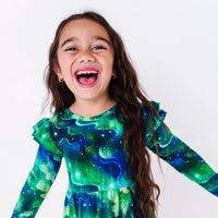 Close up portrait of a child laughing with arms out, in a long sleeve twirl dress with flutter ruffles at the shoulders in a green and blue northern lights print with tiny stars.